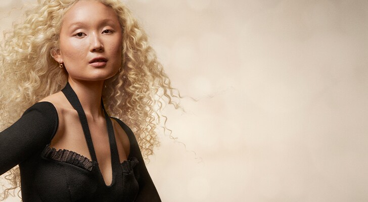 asian model with blonde wavy hair in a black top on a gold shimmer background